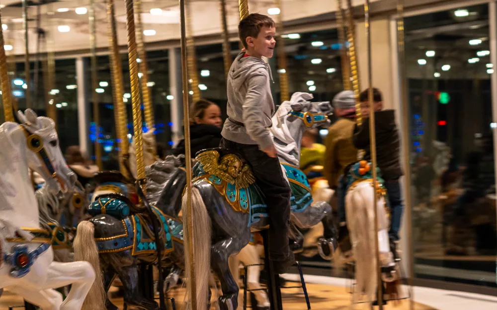 Person riding carousel horse with city lights visible through windows in background, motion blur effect