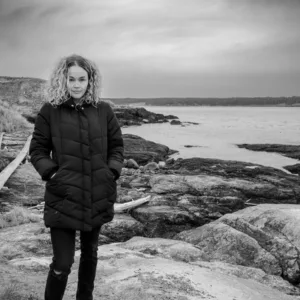 Woman in black jacket standing on rocky coastline with water and hills in background, black and white photograph