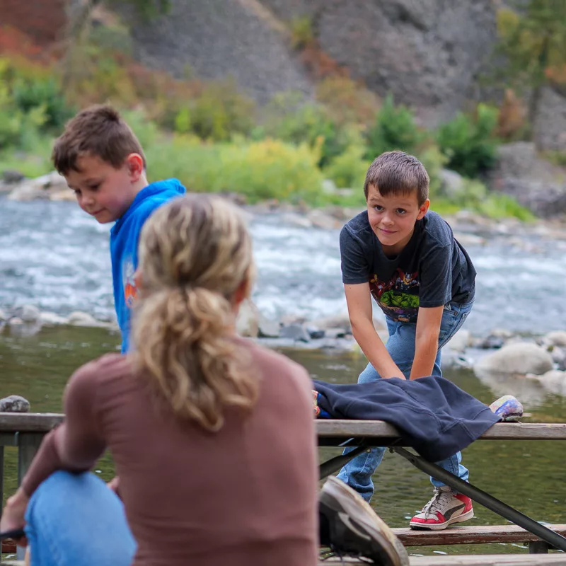 Woman sitting on bench watching two young boys playing by river with mountains in background