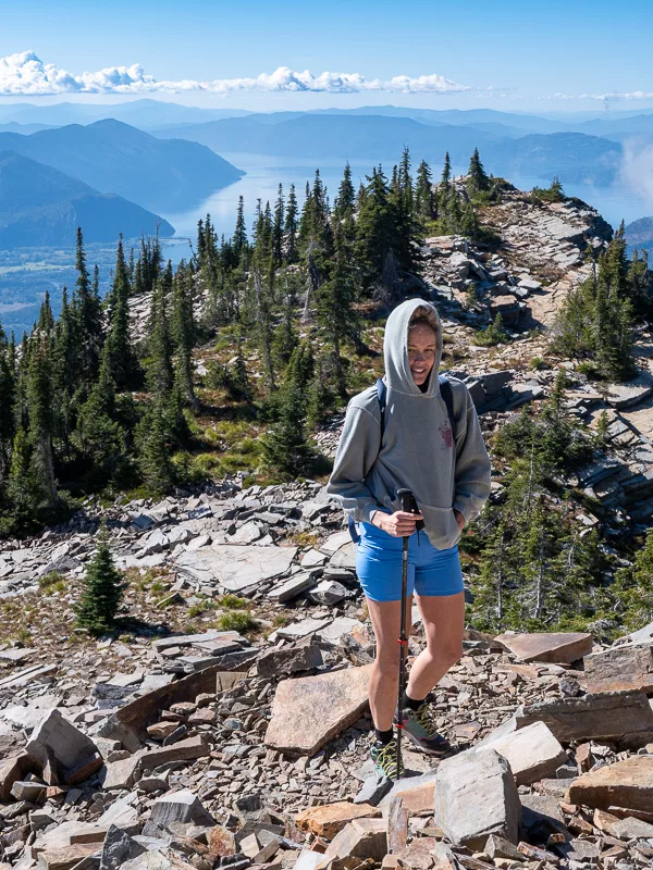 Woman in gray hoodie standing on rocky mountain trail with sweeping mountain vista behind her