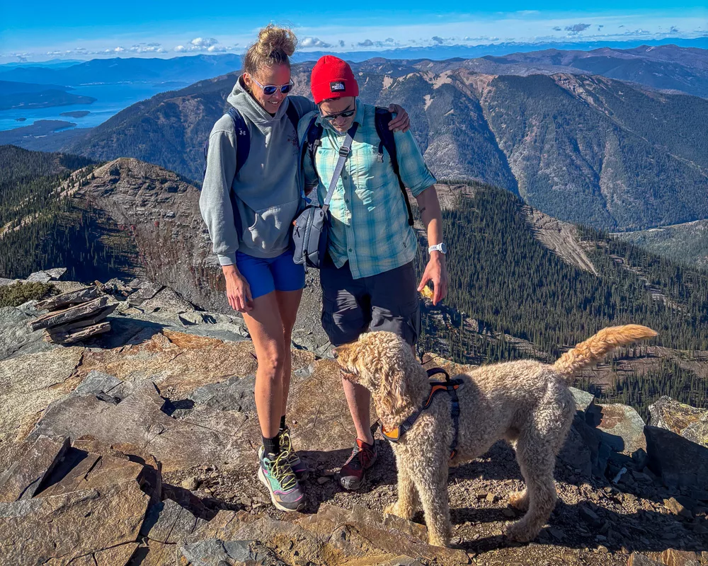 Woman in gray hoodie standing on rocky mountain trail with sweeping mountain vista behind her