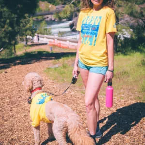 Erika Solberg in a yellow 2026 Bloomsday finisher shirt walking a trail above the Spokane River with her goldendoodle Freddy in a matching yellow shirt