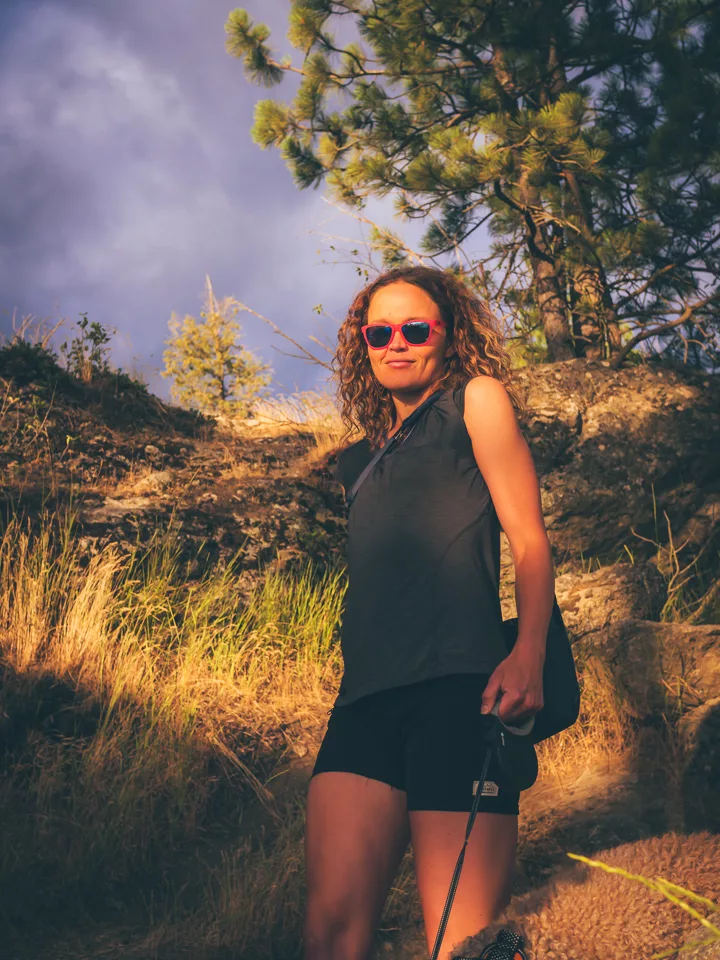 Erika Solberg standing on a rocky hillside trail at golden hour, backlit by low sun with a stormy sky and ponderosa pines behind her