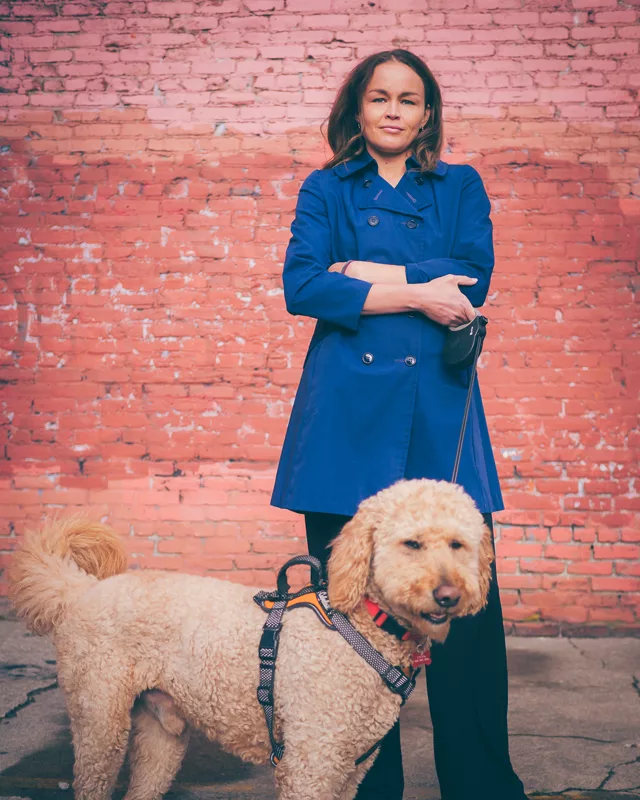 Erika Solberg standing with her arms crossed against a weathered red brick wall, with her goldendoodle Freddy on a leash beside her