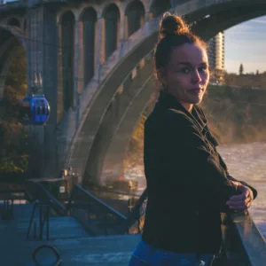 Erika Solberg stands at the Spokane River walk at golden hour, looking back over her shoulder with the Monroe Street Bridge and Gondola Skyride in the background — nine months sober, April 2026