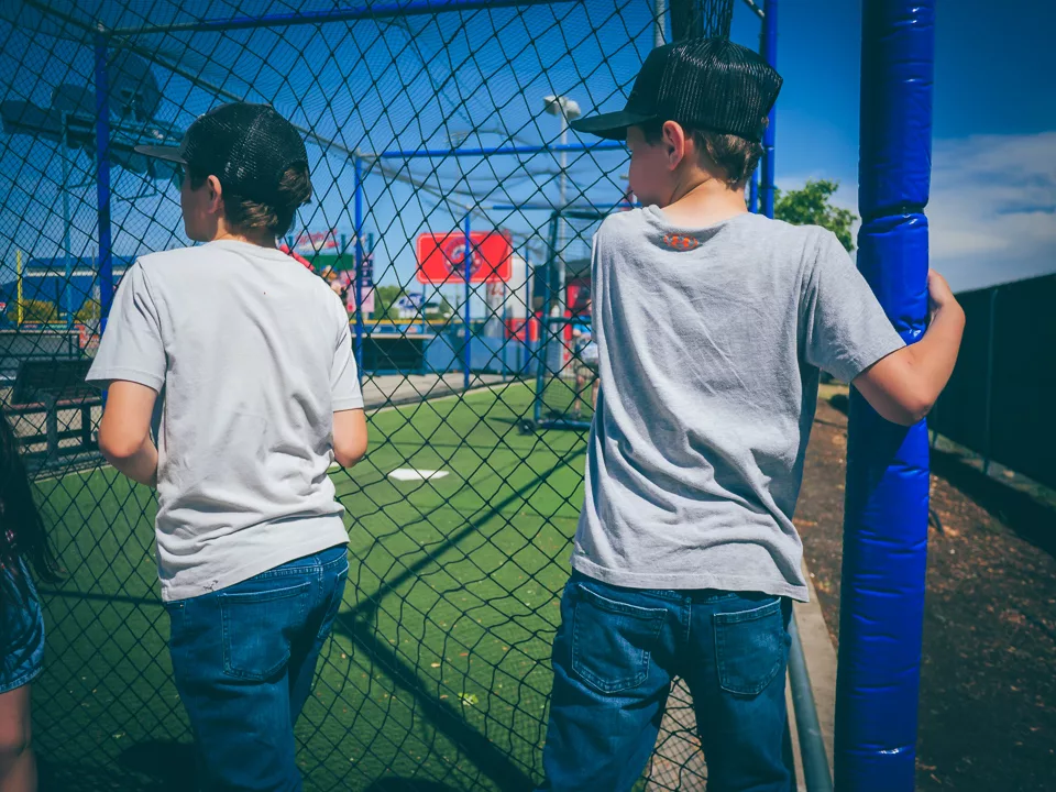 Two boys in ball caps stand with their backs to the camera at a chain-link batting cage on a sunny day