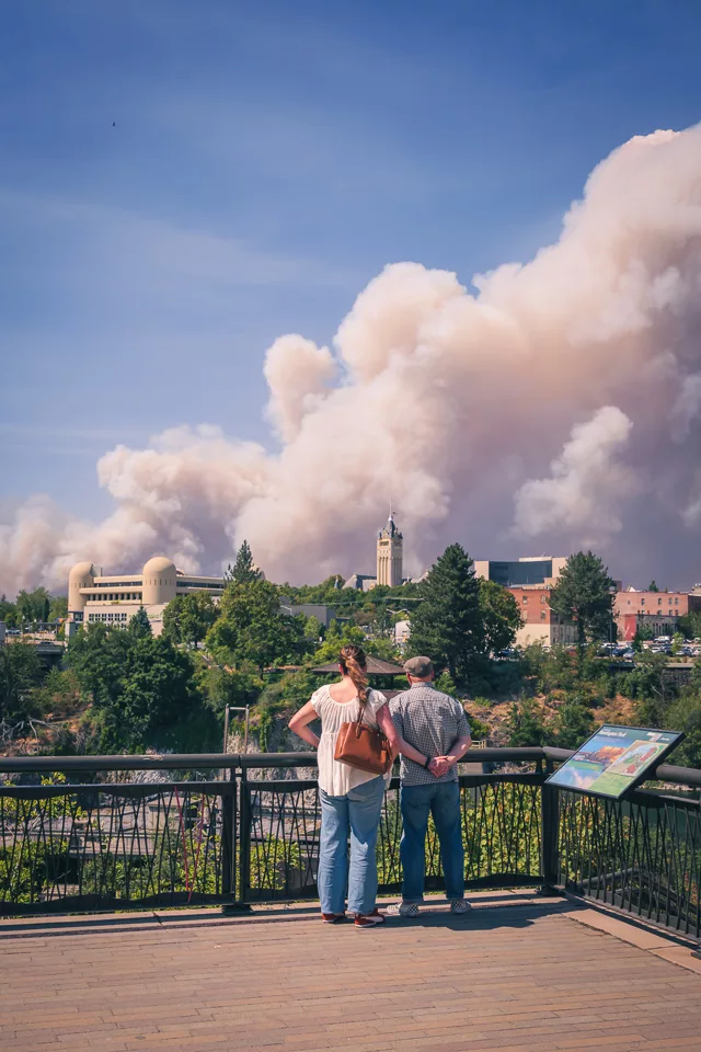 A couple stand at a downtown Spokane overlook watching a towering smoke plume rise behind the county courthouse clock tower